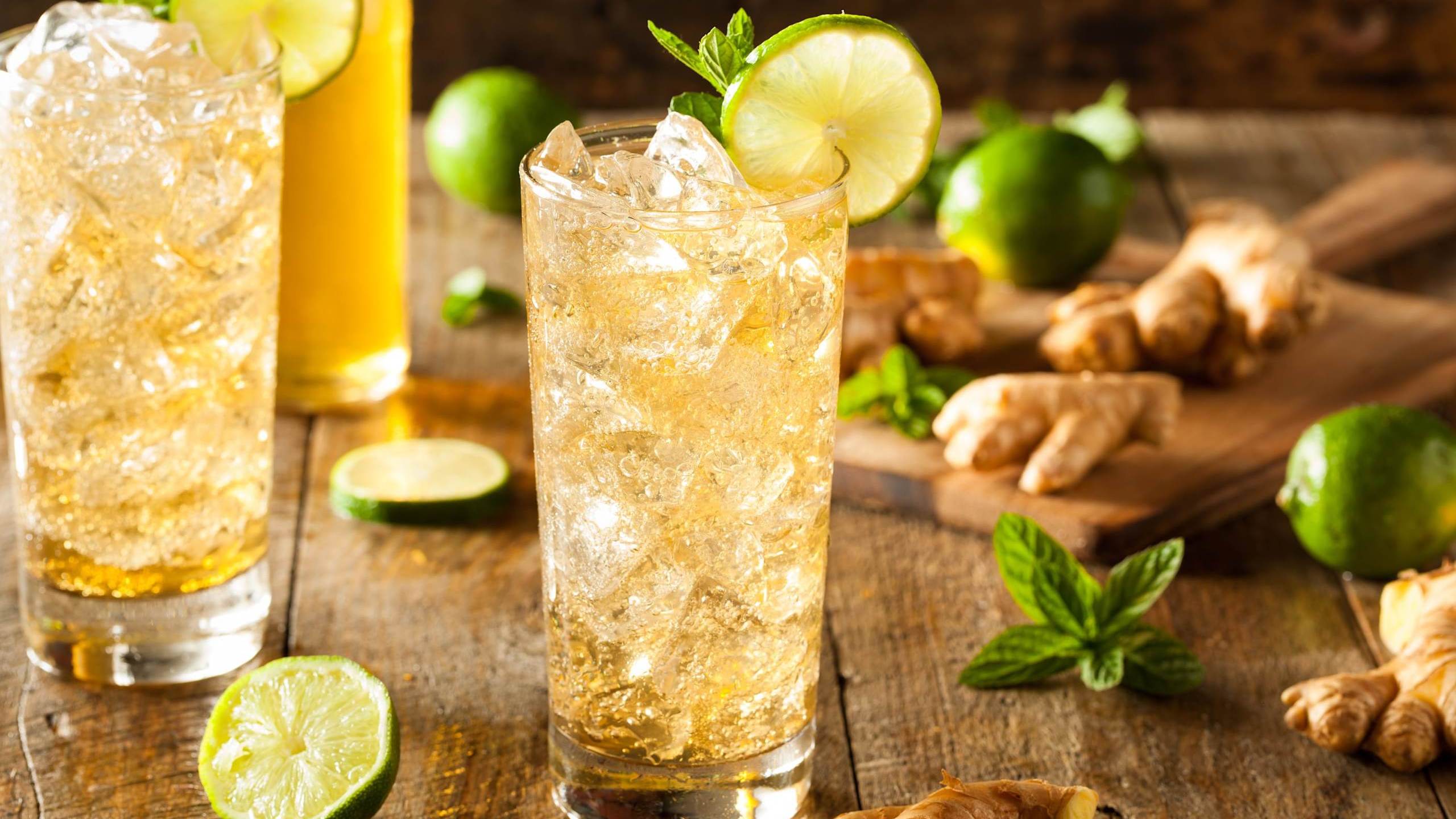 Deux grands verres remplies de glaçons avec une boisson sans alcool dorée et pétillante. Des racines de gingembre, des tranches de lime et des feuilles de menthe sont parsemés autours.