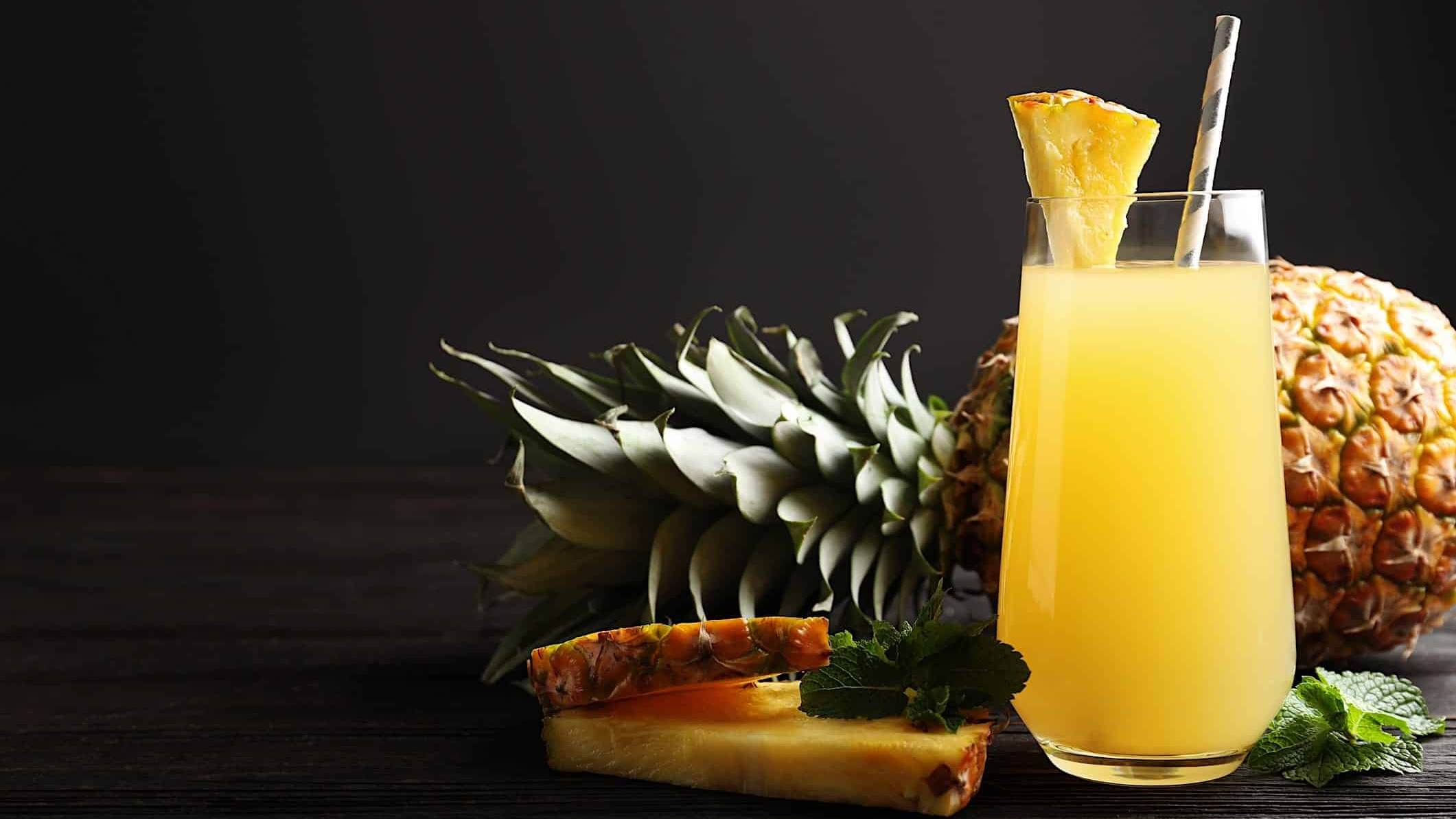 Un grand verre d'une boisson sans alcool à l'ananas entouré d'un ananas entier et de quelques tranches garnies de menthe