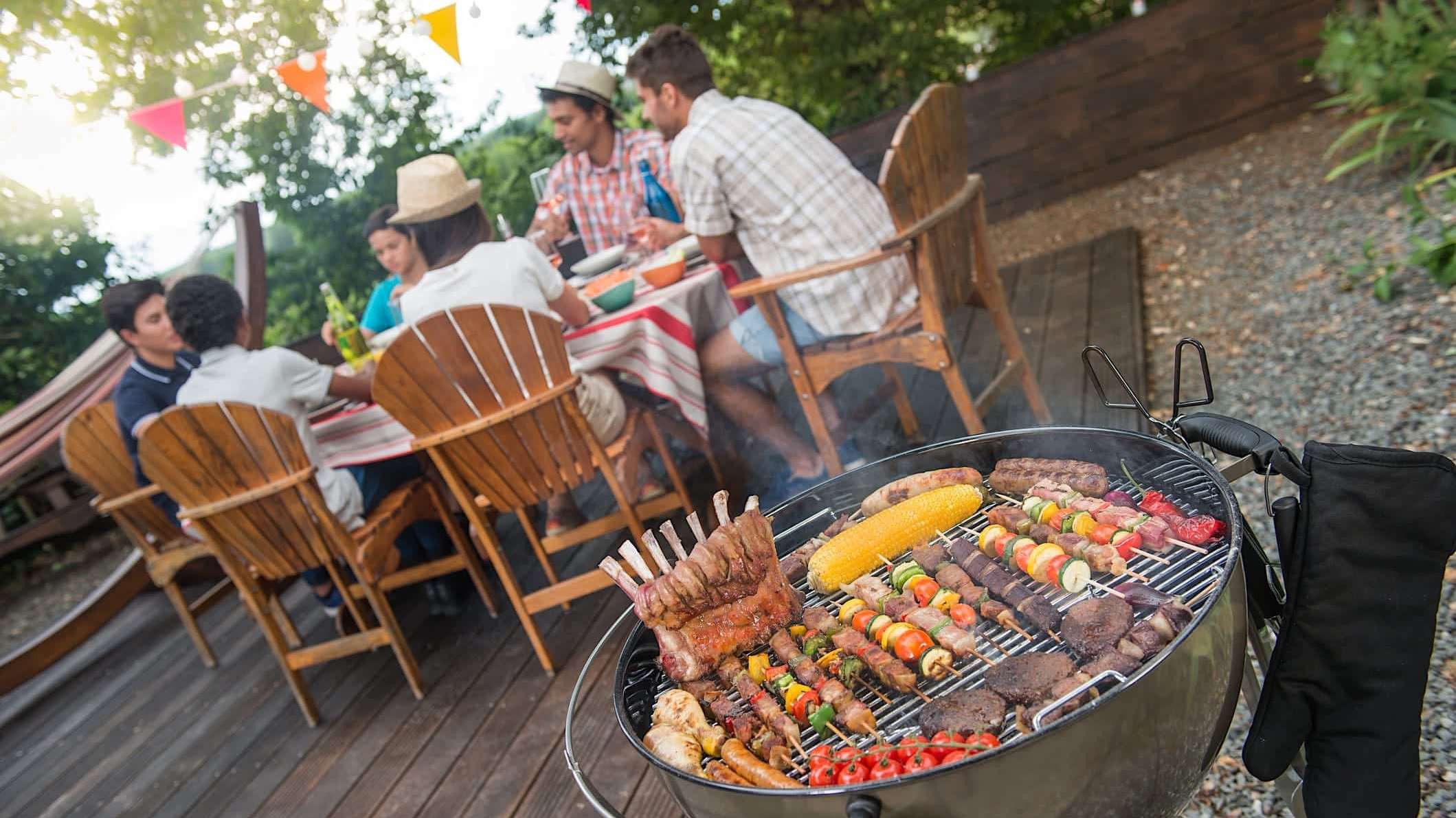 Gros plan sur un BBQ au charbon où grillent des viandes et légumes, avec une famille à table en arrière plan