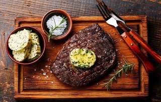 Un steak grillé au BBQ garni d'une tranche fondante de beurre composé est posé sur une planche de bois. Un bol avec quelques tranches froides de beurre composé y est posé, avec un ramequin de gros sel et une paire d'ustensile.
