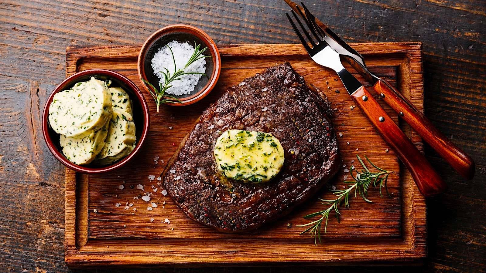 Un steak grillé au BBQ garni d'une tranche fondante de beurre composé est posé sur une planche de bois. Un bol avec quelques tranches froides de beurre composé y est posé, avec un ramequin de gros sel et une paire d'ustensile.