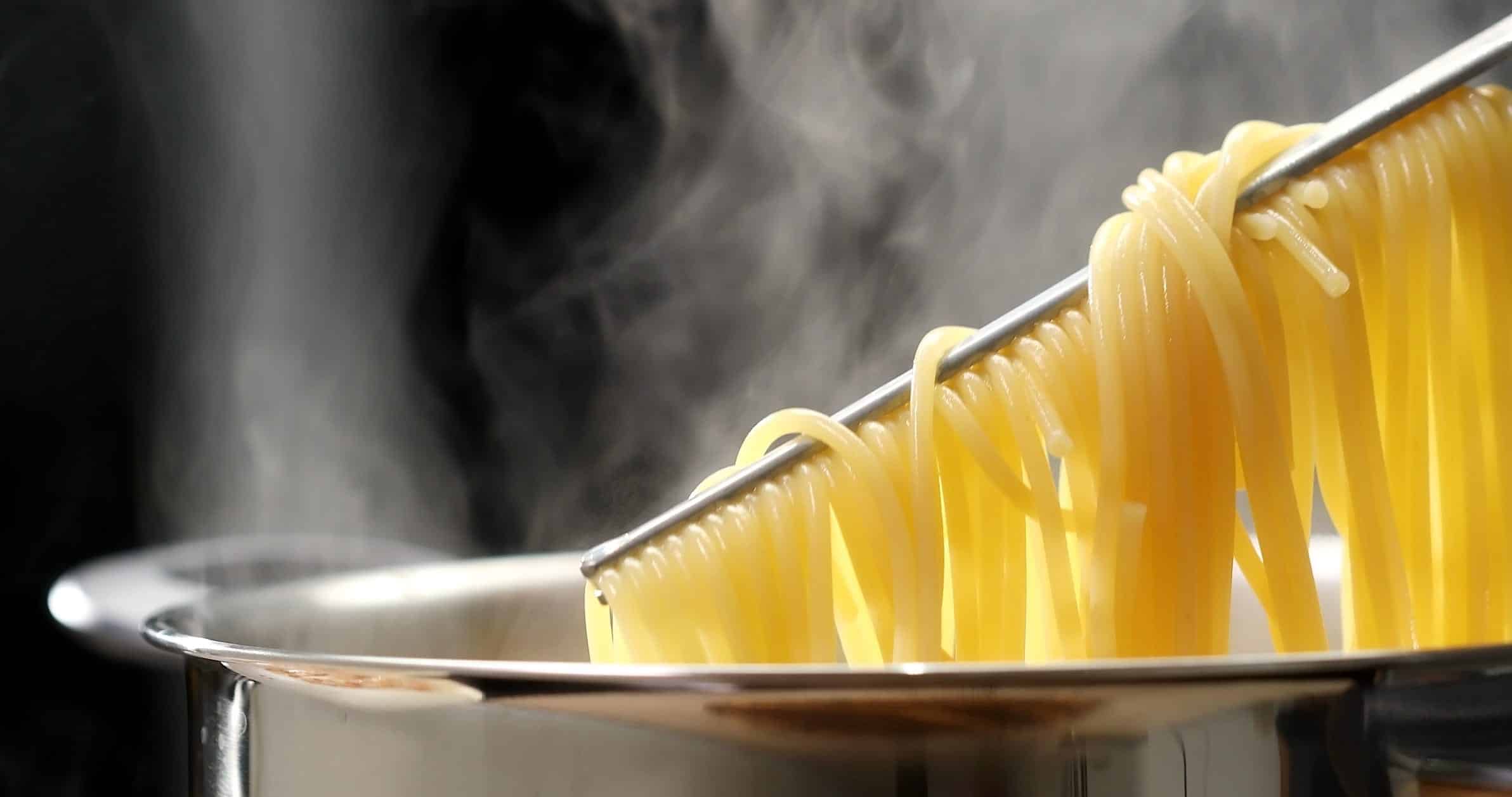 Des spaghettis sont soulevés avec une pince d'une casserole qui émet de la vapeur.