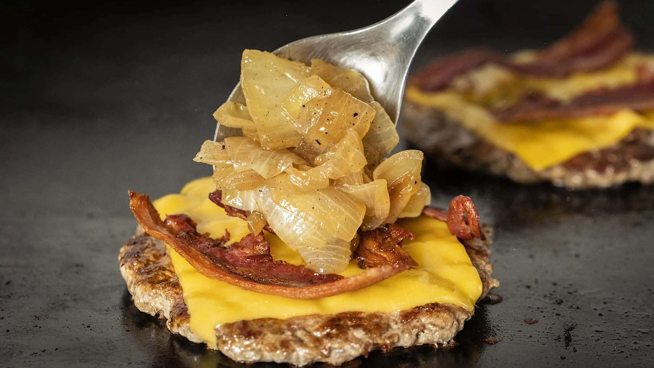 Une boulette de burger cuit sur une plancha BBQ, garnie de fromage en tranche, de bacon et d'oignons caramélisés