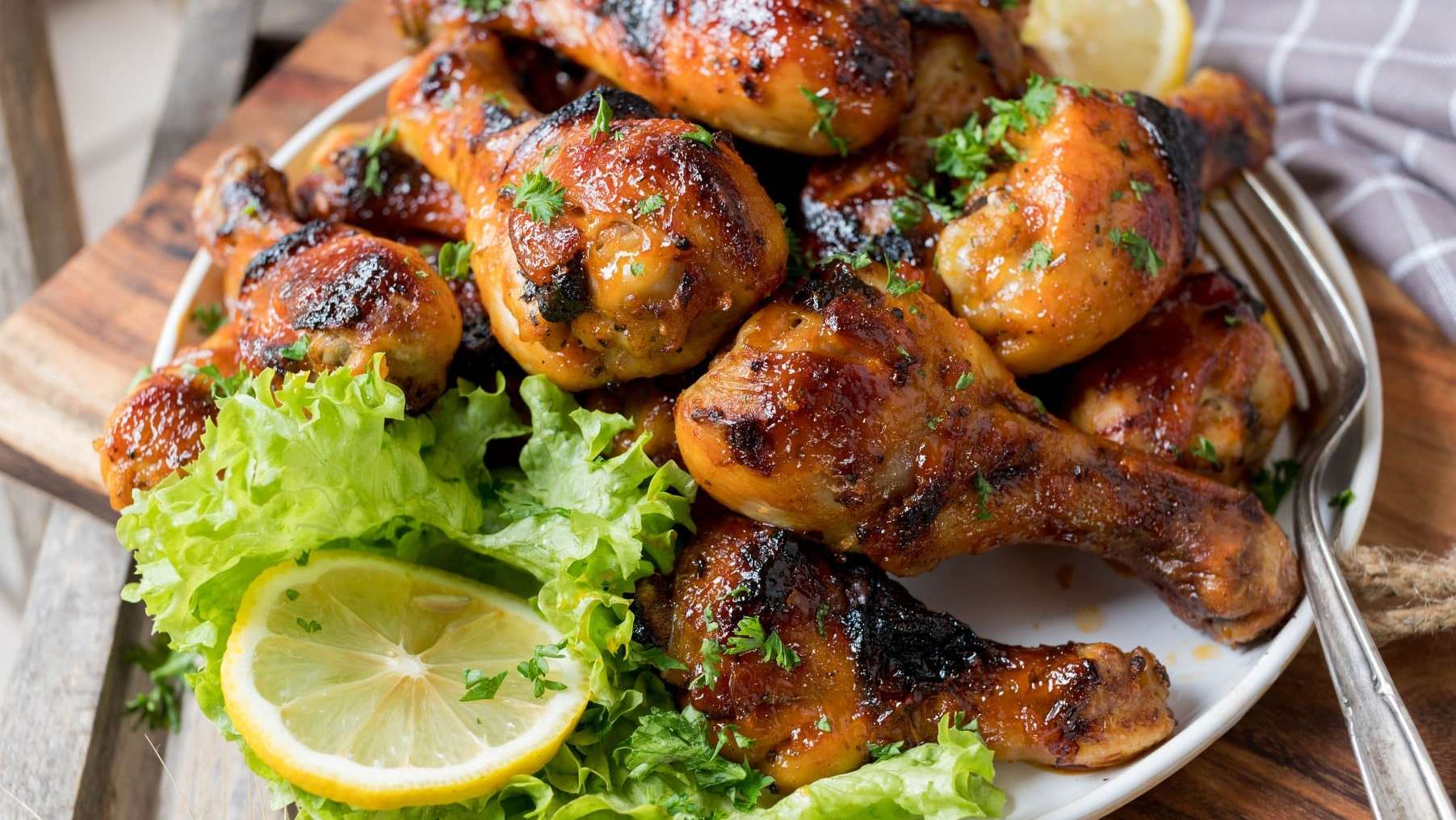 Des pilons de poulet marinés et cuits au BBQ sont posés dans une assiette blanche et garnis de laitue frisée et de tranches de citron