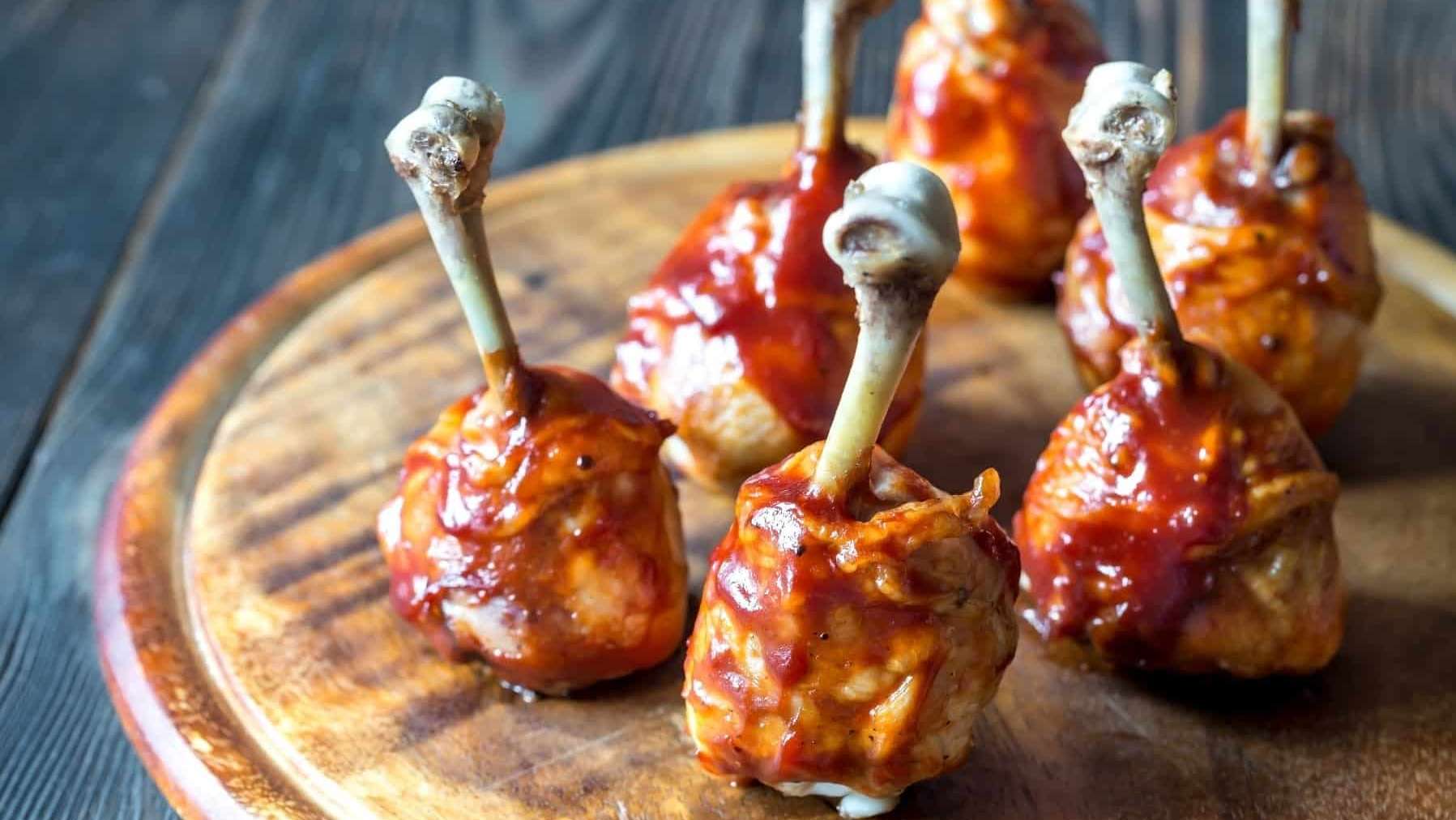 Des lollipops de pilons de poulet marinés et cuits au BBQ sont posés sur une planche de bois ronde.