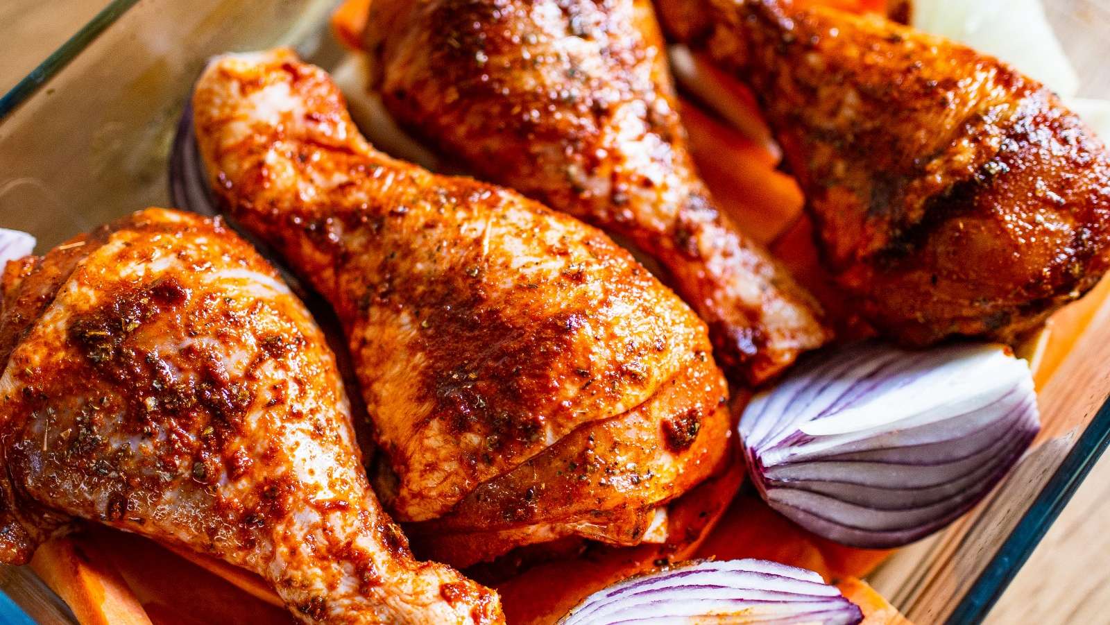 Des pilons de poulet marinés pour une cuisson au BBQ sont posés dans une casserole rectangulaire en pyrex avec des quartiers d'oignon rouge.