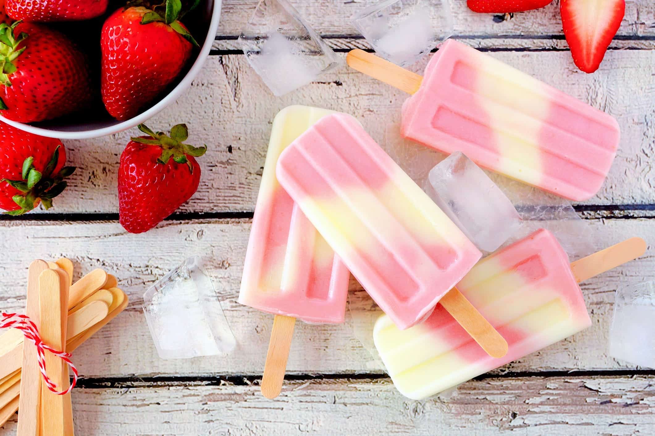 Fruits-surgeles-recettes-bbq-popsicle-fraise-creme 4 popsicles fraises et crème faits avec des fruits surgelés sont posés sur une table de bois et sont entourés de glaçons et de fraises fraîches