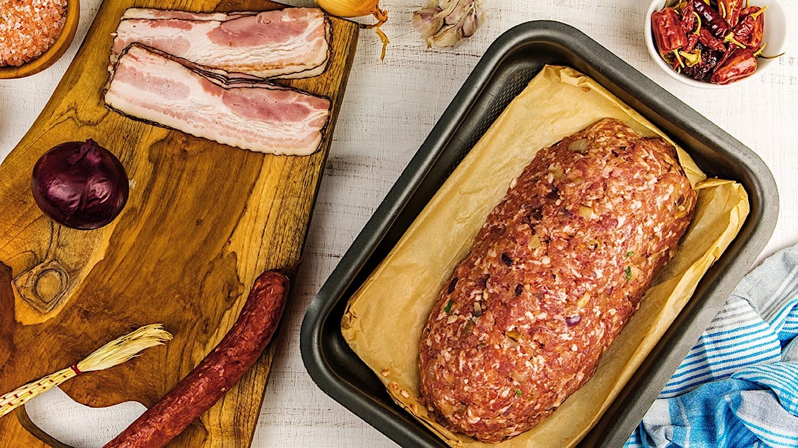 Un pain de viande placé sur une plaque de cuisson, accompagné de quelques ingrédiens posés sur une planche à découper