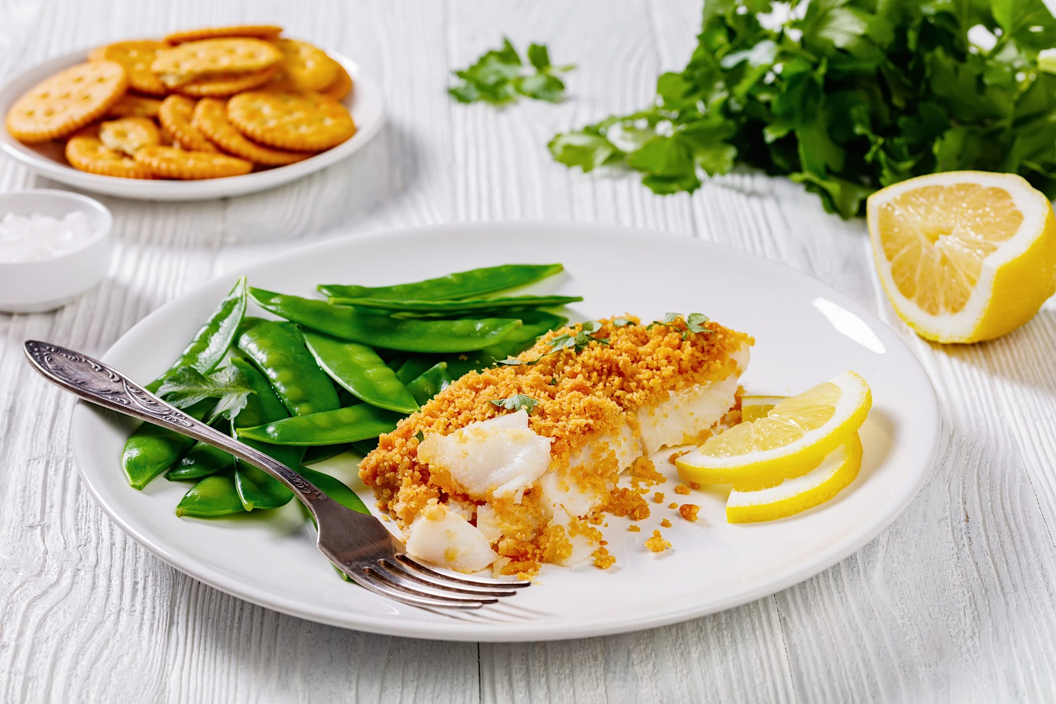 Cuisson-poissons-blancs-recettes-gratine un filet de poisson blanc garni de chapeleure gratinée est dressé dans une assiette blanche et accompagné de pois mange-tout et de tranches de citron.
