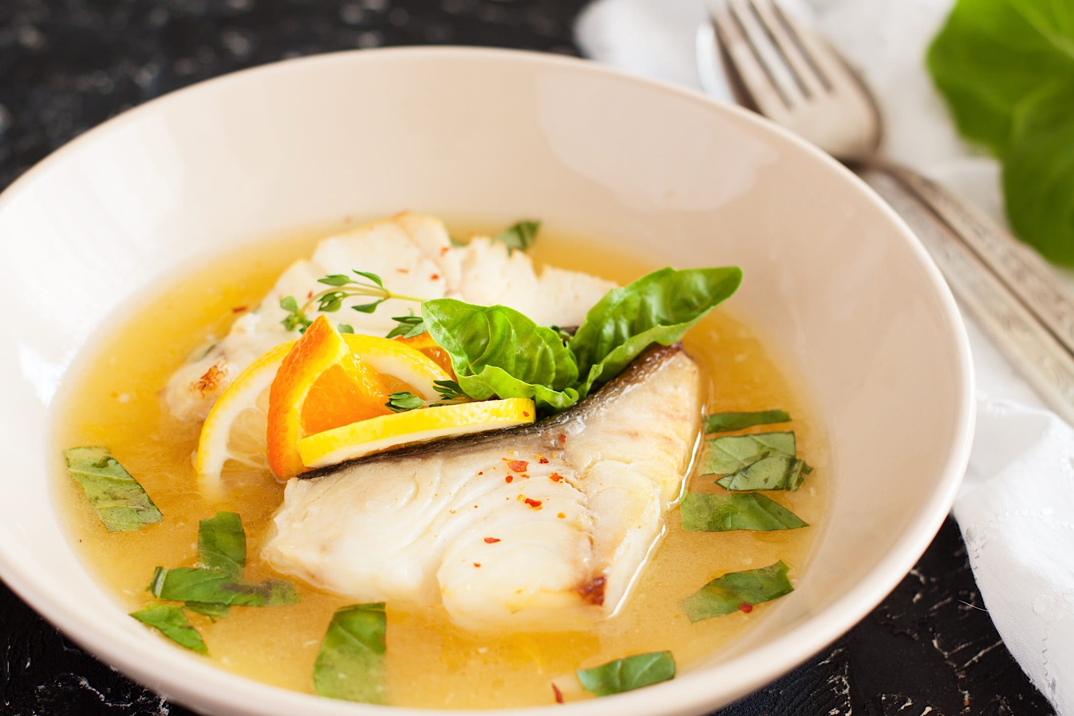 Cuisson-poissons-blancs-recettes-poche-vapeur Un filet de poisson blanc poché ou cuit vapeur est posé dans un bol blanc contenant un bouillon garni d'herbes fraîches et de tranches d'orange.