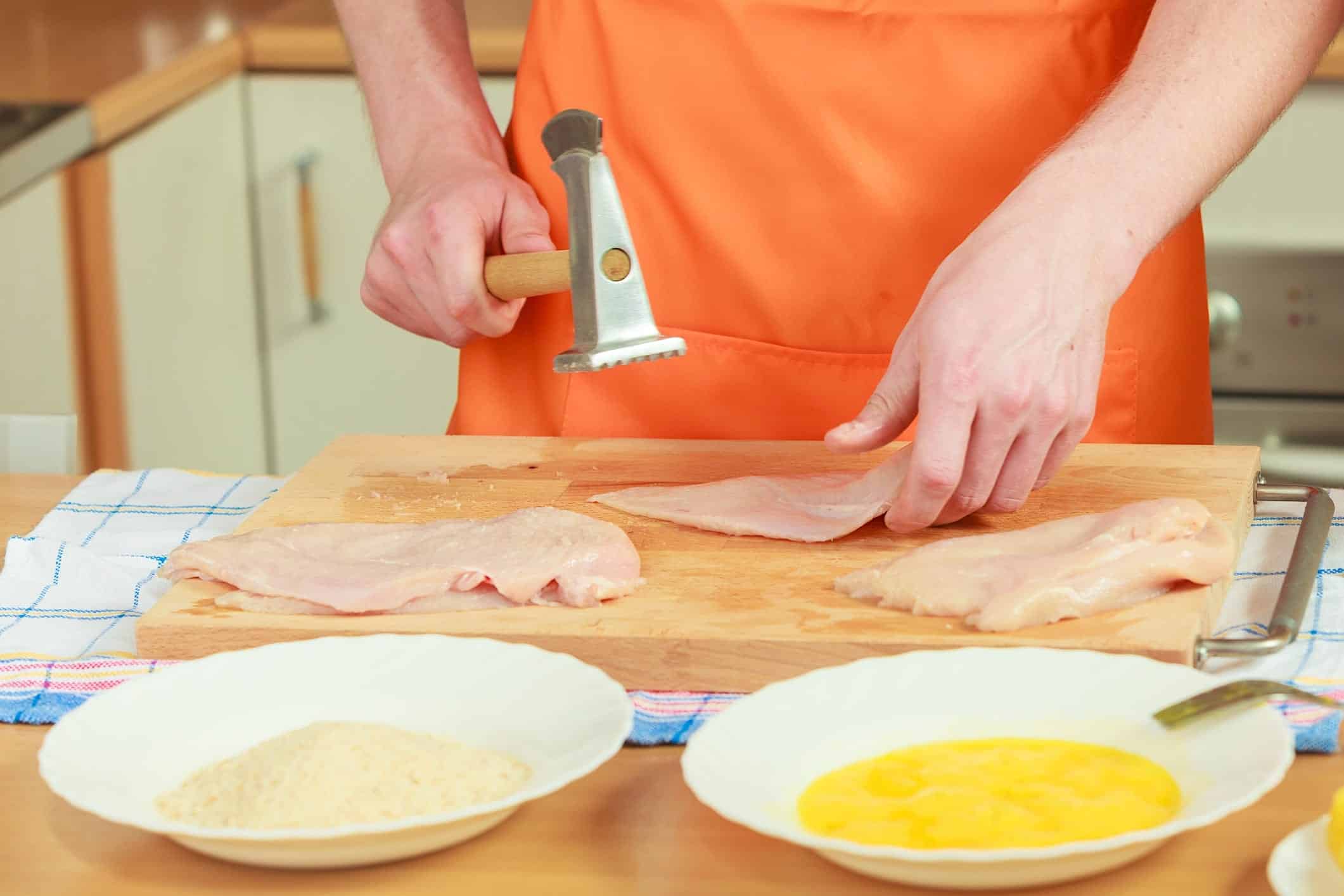 Poulet-parmesan-recette-paner La main d'une personne tient un marteau à viande au dessus d'une escalope de poulet cru posée sur une planche, accompagné d'un bol de chapelure et d'un bol d'oeufs fouettés pour faire une panure à l'anglaise.