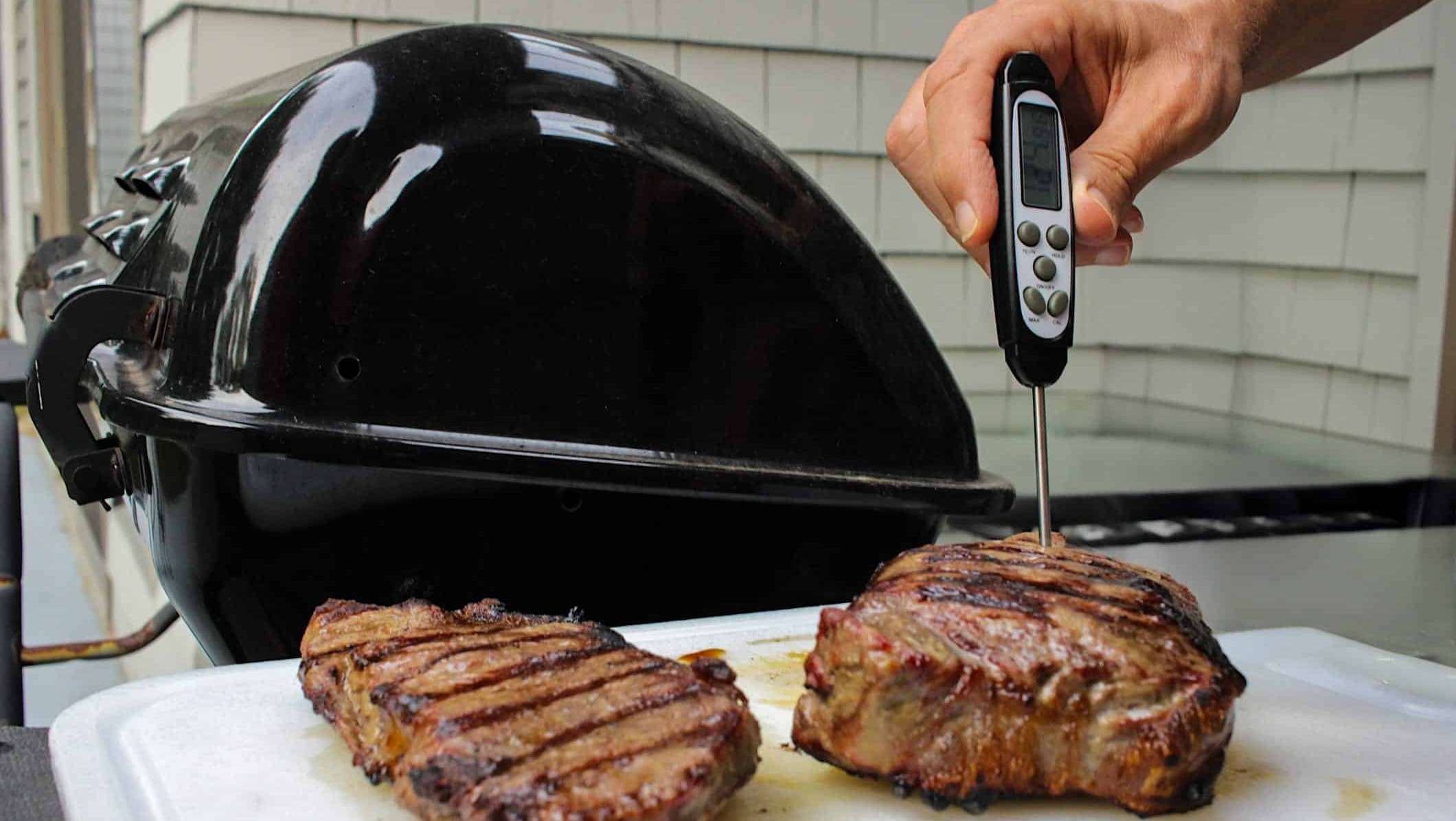 Cuisson-steak-BBQ-thermometre-temperature-interne Deux steaks sont posées sur une planche découper noire posée à côté d'un BBQ, et la main d'un homme prend la température interne du steak avec un thermomètre à lecture instantanée.