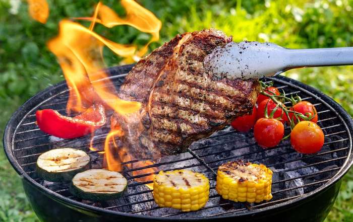 Une personne tient un steak grillé avec des pinces au dessus d'un BBQ au charbon recouvert des légumes grillés