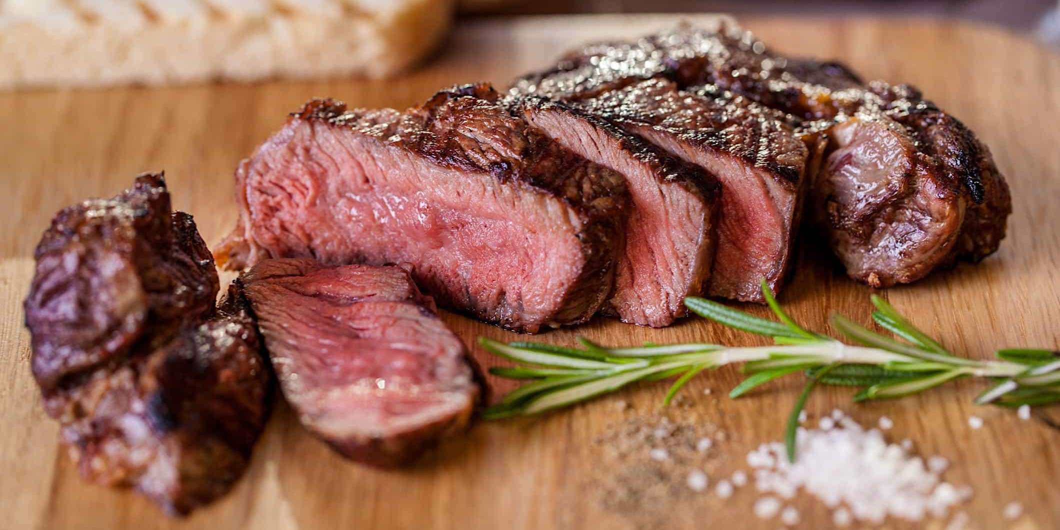 Cuisson-steaks-BBQ-cuit Un steak avec une cuisson parfaite, grillé au BBQ, est tranché et placé sur un plateau en bois, garni d'une branche de Romarin et d'un tas de sel de mer.