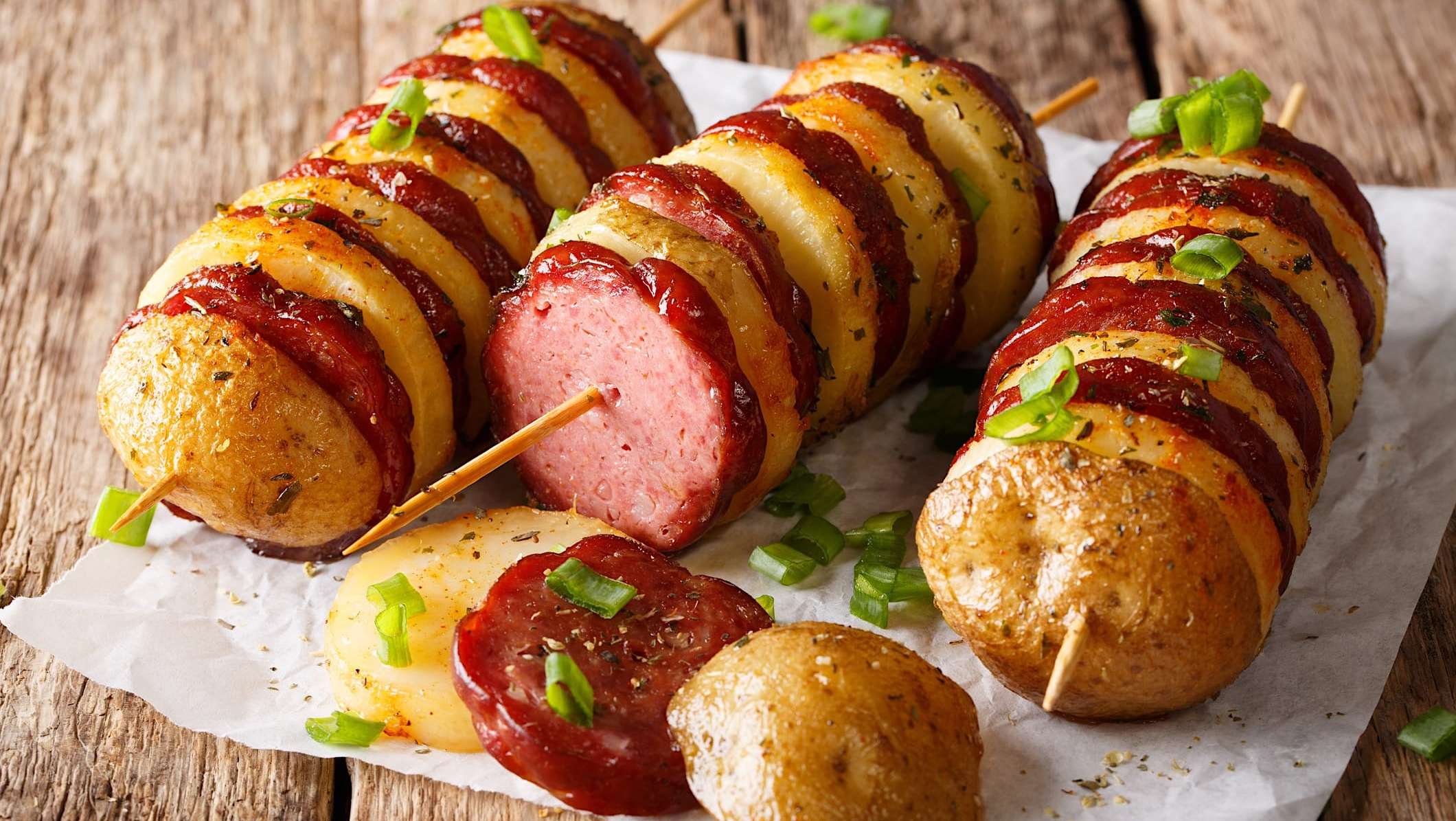 recettes-pommes-de-terre-BBQ-brochette-saucisse Des brochettes de tranches de pommes de terre et de tranches de saucisses grillées au BBQ sont posées sur un papier parchemin, sur une table en bois.