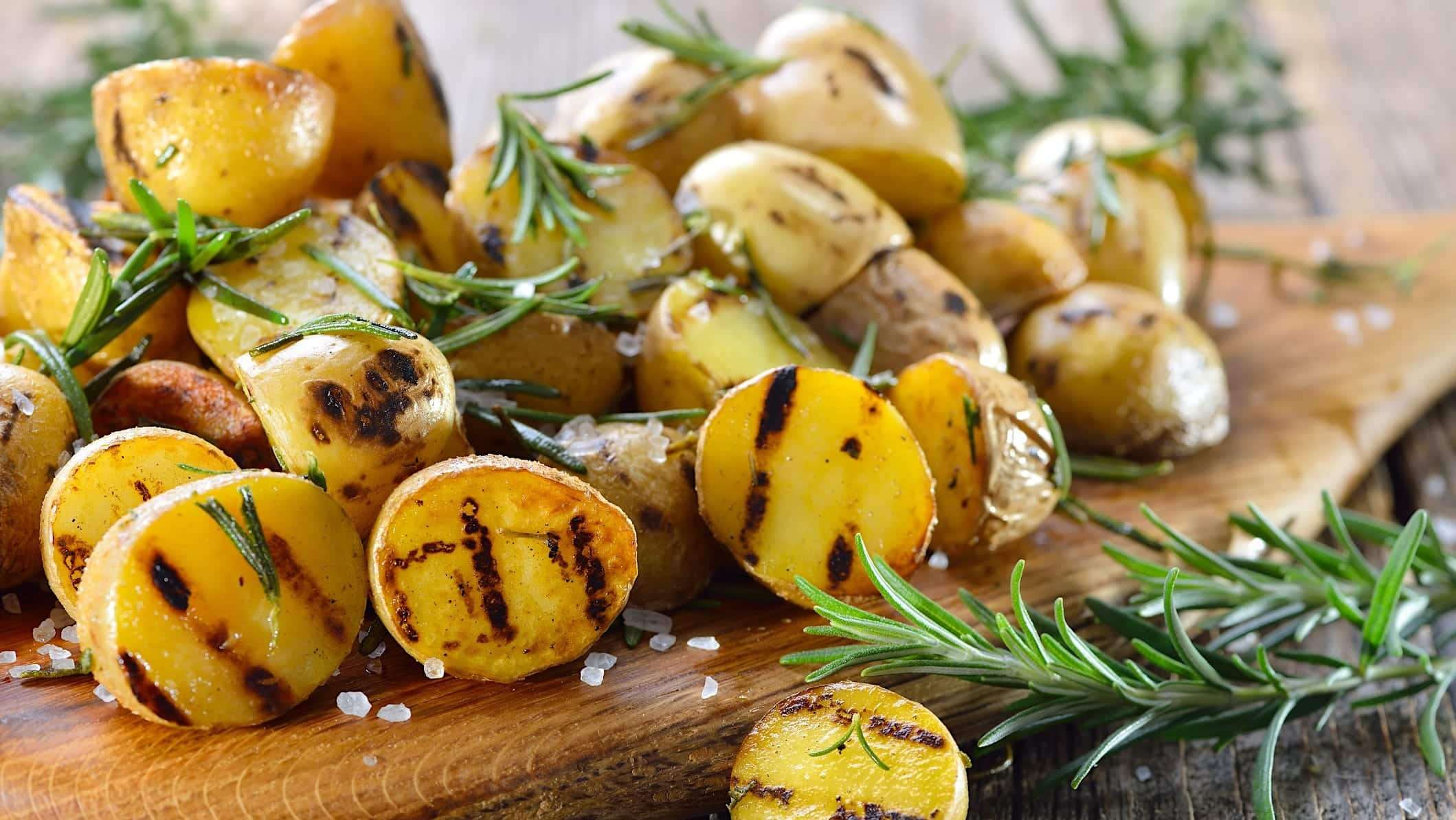 recettes-pommes-de-terre-BBQ-grillées Une recette de pommes de terre grelots coupées en deux et grillées sur le BBQ sont posées sur une planche de bois avec du romarin frais