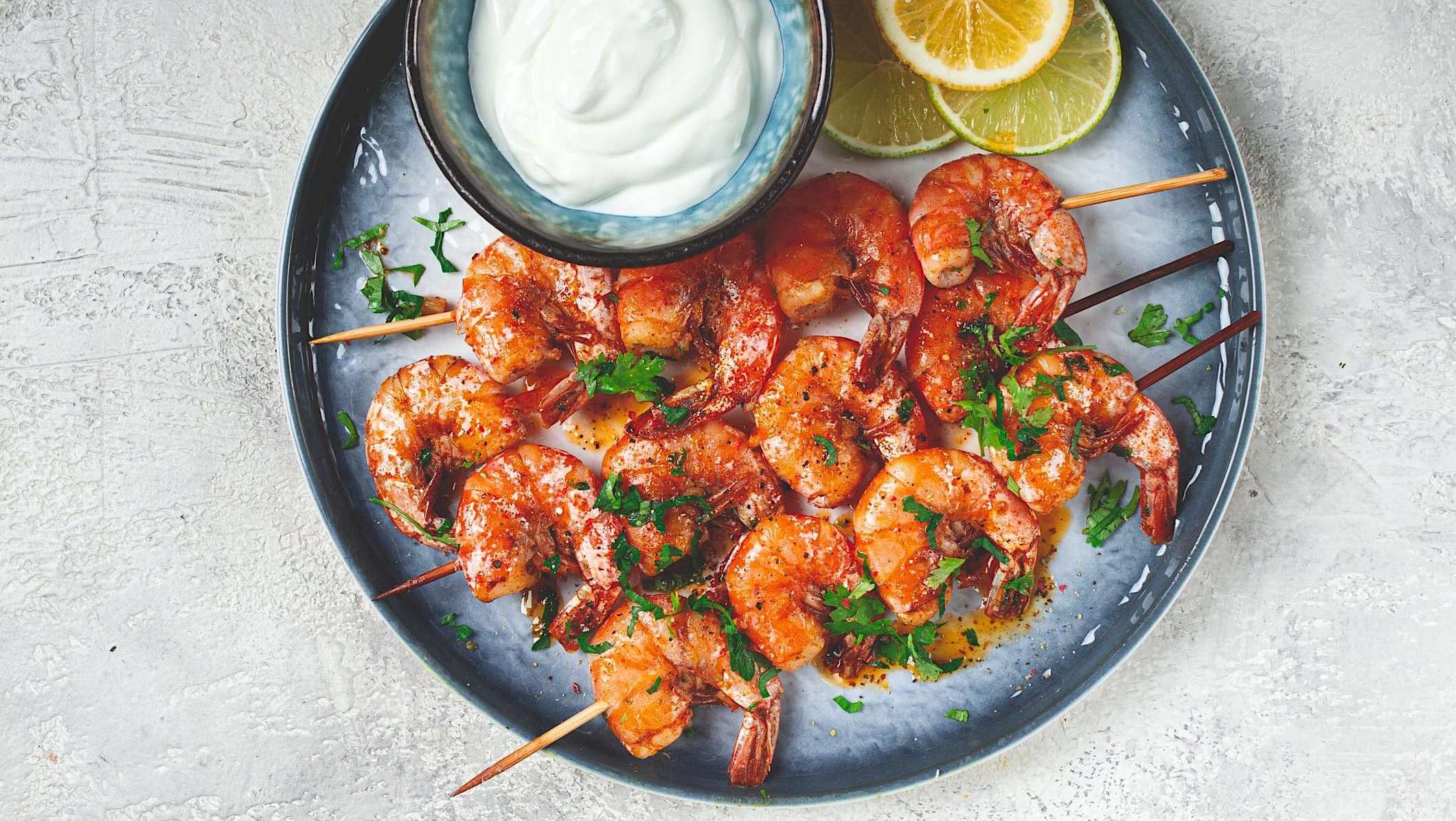 Une assiette bleue est posées sur un comptoir de marbre, et 3 brochettes de crevettes grillées au BBQ y sont dressées avec un bol de sauce et quelques tranches d'agrumes.