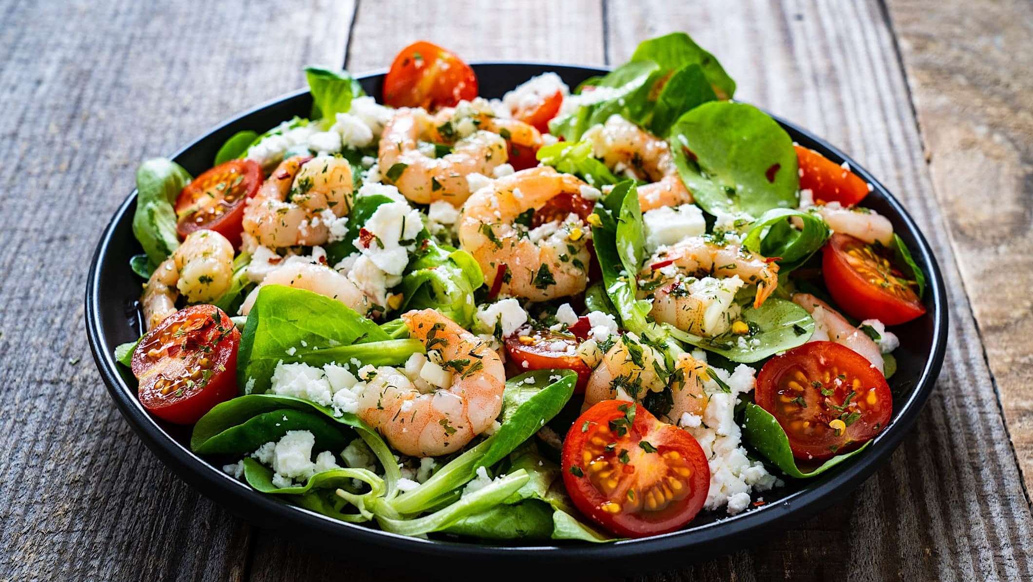 Une salade contenant des crevettes marinées et grillées au BBQ, avec des tomates cerise, des épinards et du fromage feta.