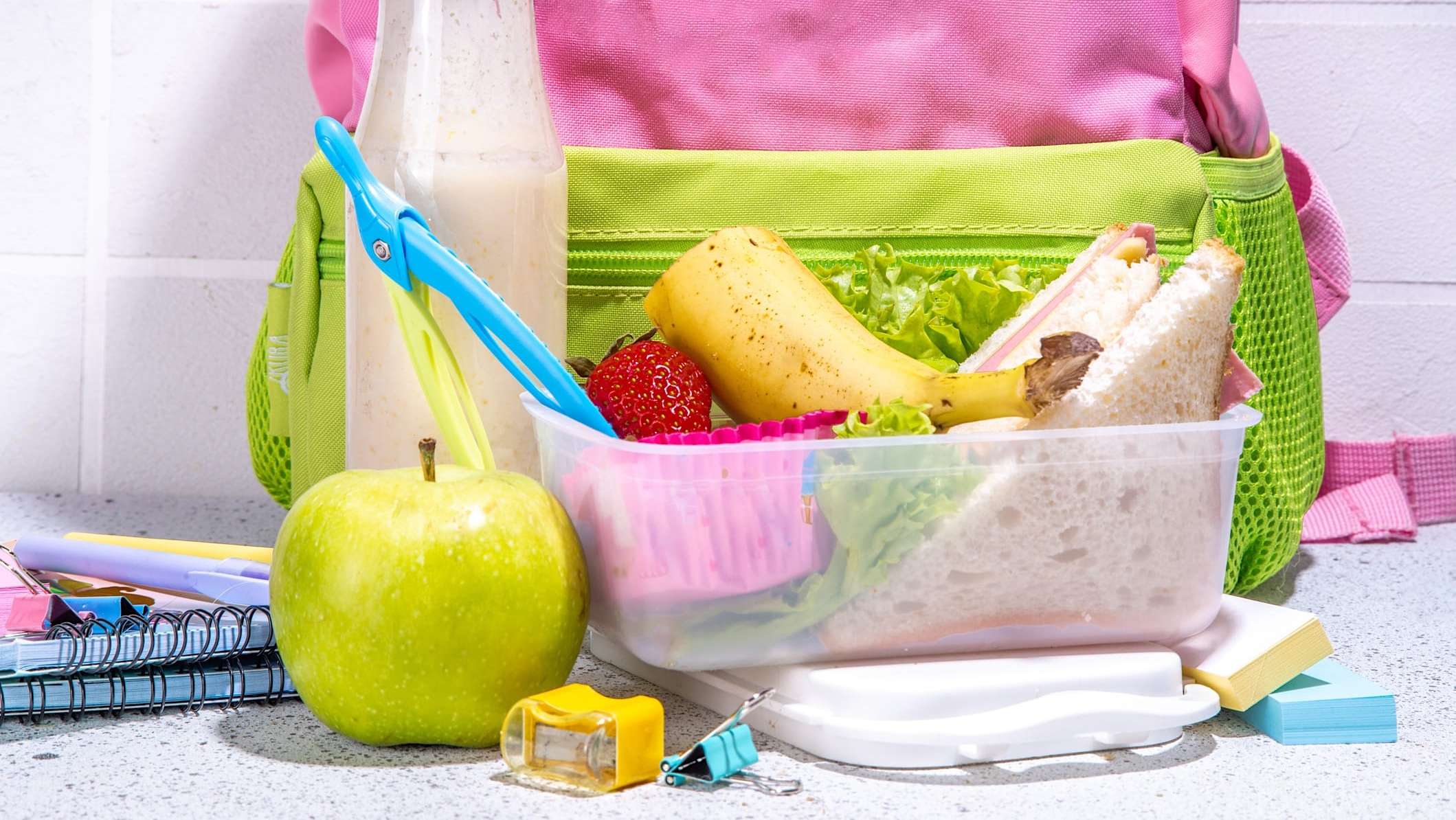 enfant-difficile-nourriture-boite-lunch Devant un sac à dos d'enfant, une boîte à lunch comportant un demi sandwich au jambon, des fraises et une demie banane, accompagnée d'une pomme verte.
