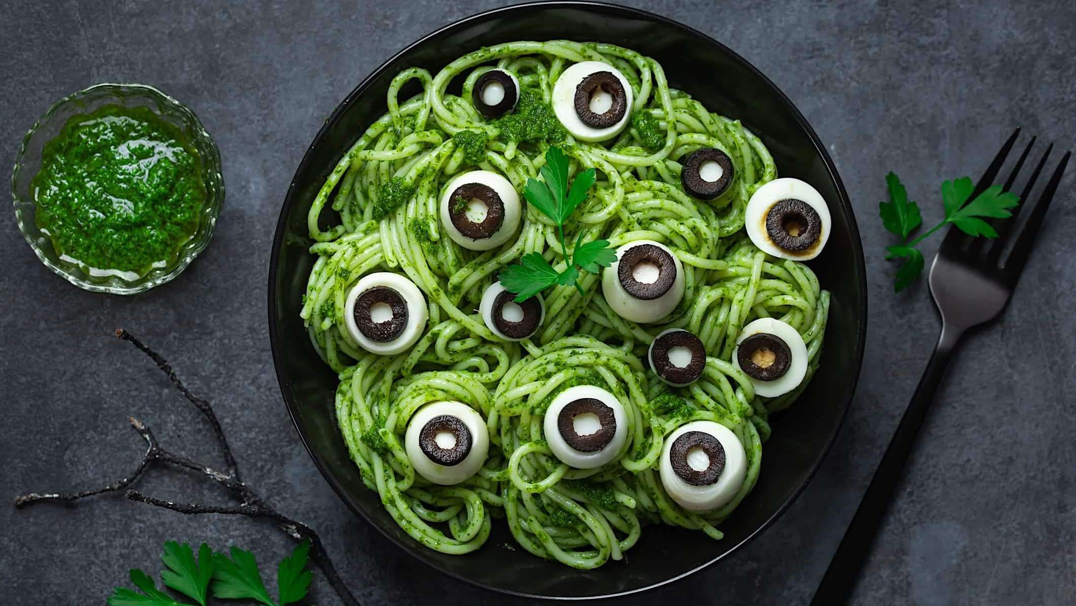 boite-lunch-halloween-spaghetti-pesto-yeux Une assiette noire contient des spaghettis au pesto de basilic avec des boules de bocconcini décorés comme des yeux avec des tranches d'olive noire pour une boite à lunch de l'Halloween