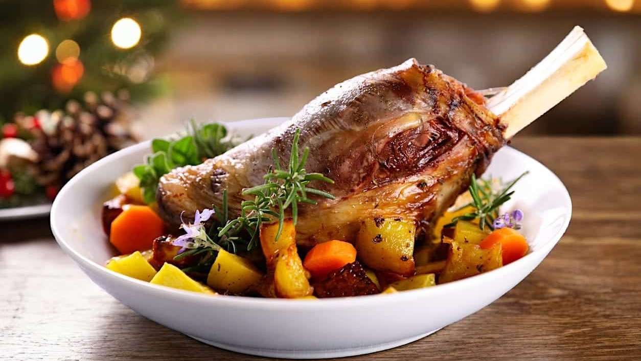 Un gigot d'agneau braisé et rôti est posée dans un grand bol rempli de légumes rôtis pour illustrer des idées de repas pour remplacer la classique dinde de Noël.
