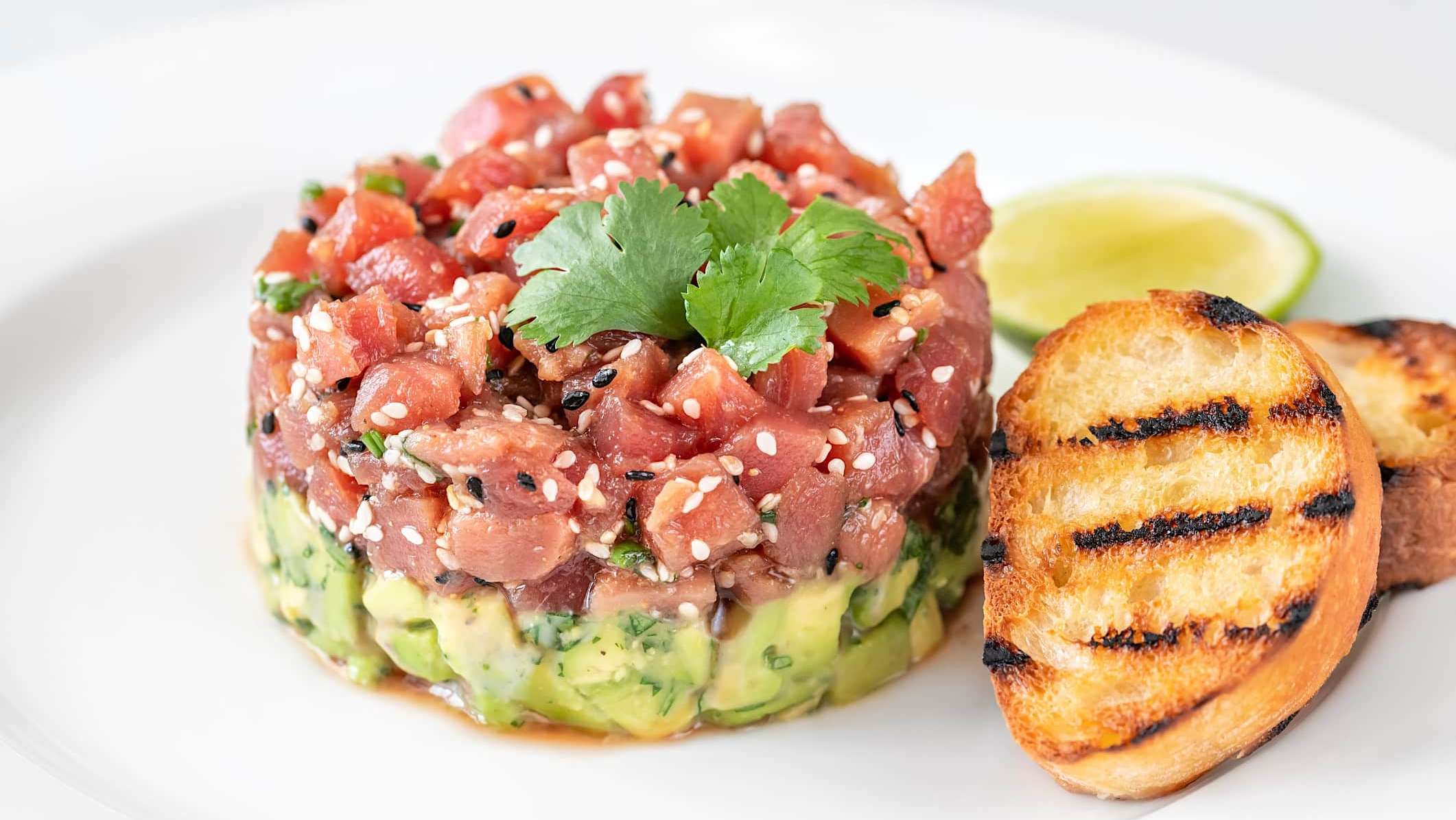 Une recette de tartare de thon à l'avocat est dressé dans une assiette et accompagné de croutons de pain
