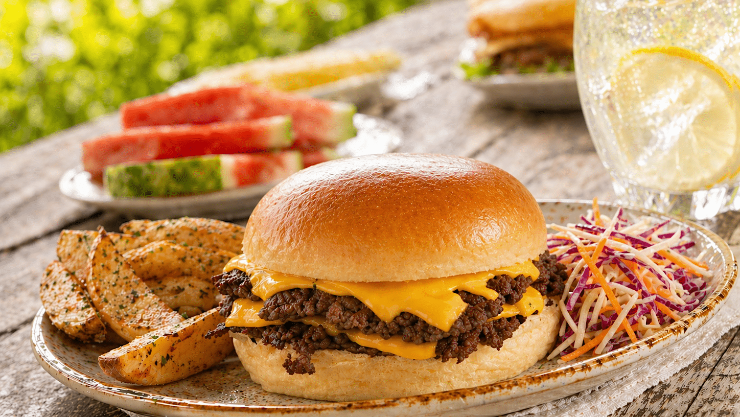 Une assiette garnie d'un double smash burger au fromage, de quartiers de pommes de terre et de salade de chou est placée sur une table de pique-nique avec des tranches de melon d'eau et un verre de limonade