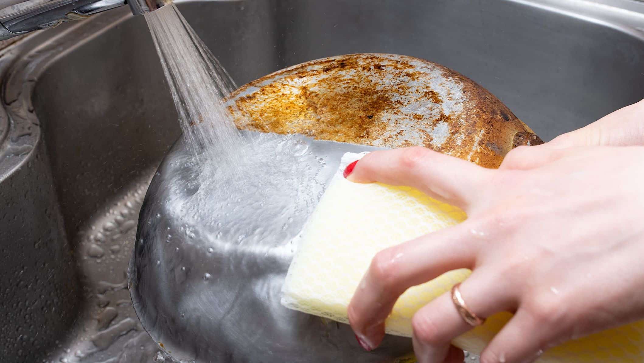 Gros plan d'une main qui nettoie avec une éponge le dessous d'une casserole en inox; une moitié est tachées et l'autre est propre, pour illustrer comment faire le grand ménage du printemps de la cuisine.